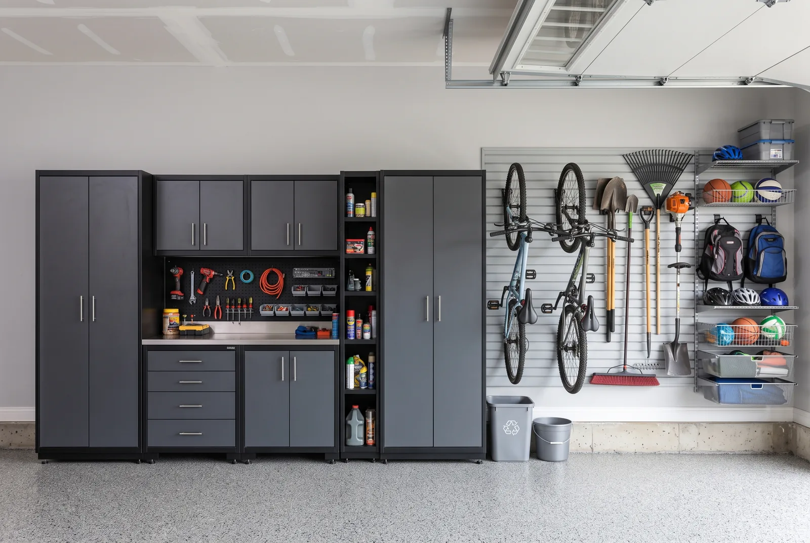 A finished garage wall: grey cabinets with a worktop and pegboard on the left, slat wall holding bikes, rakes, shovels and sports gear on the right.