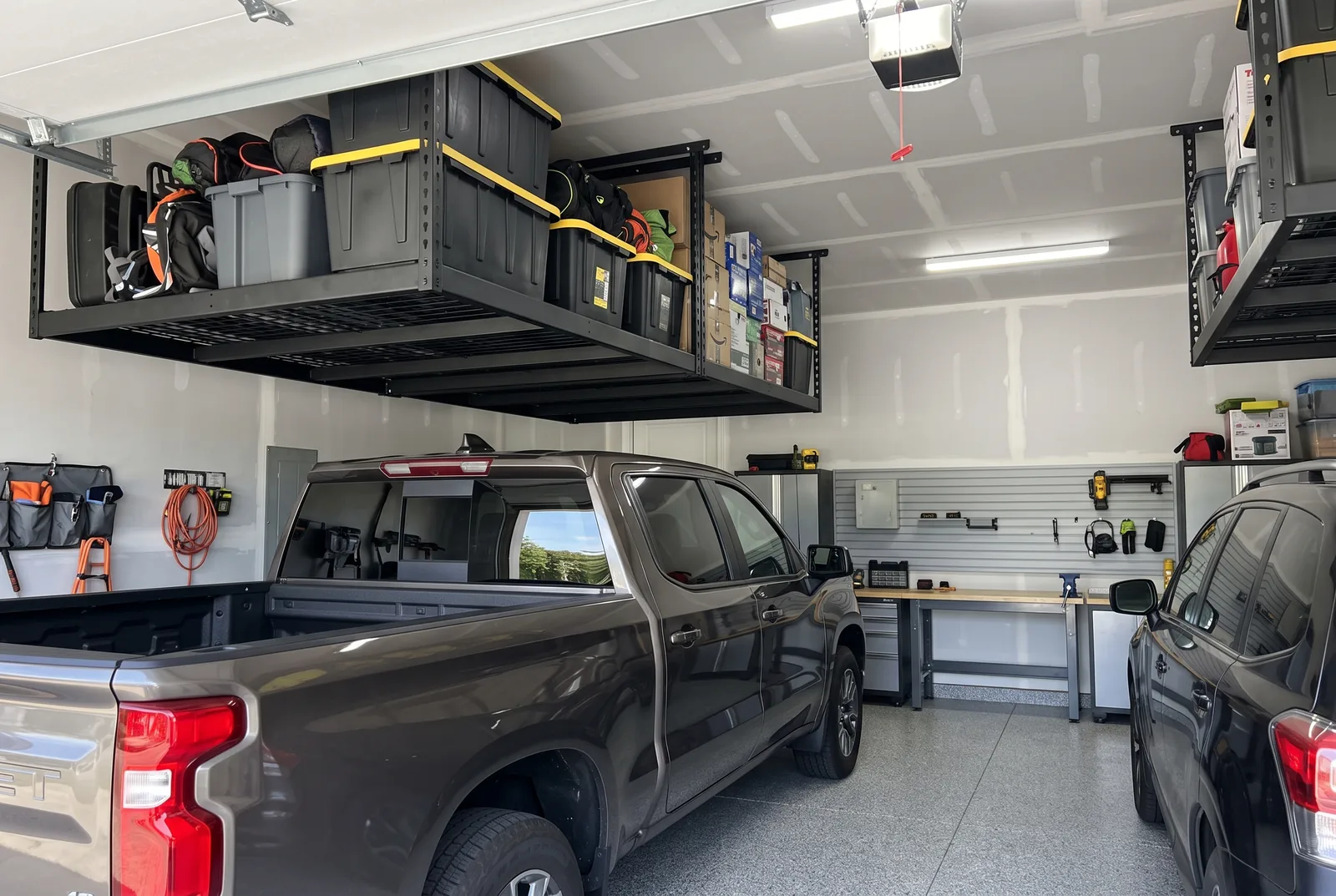 Two overhead storage racks mounted to the ceiling of a two-car garage, loaded with bins and bags, with a pickup truck parked underneath.